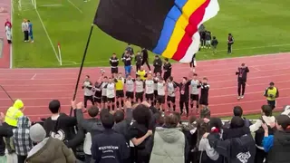 Un grup de persoane stă pe o pistă de atletism lângă un teren de fotbal, ridicând un steag mare colorat în culorile negru, albastru, galben și roșu.