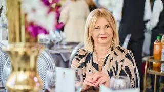 O femeie cu părul blond, purtând o bluză cu model negru pe fundal bej, stă la o masă cu pahare și obiecte de decor.
