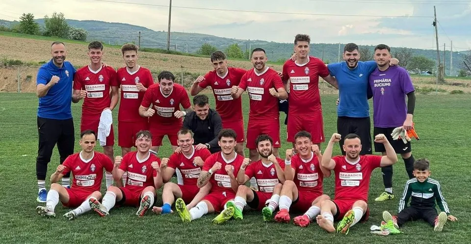 Un grup de fotbaliști în echipamente roșii și albastre pozează pe un teren de fotbal verde sub un cer înnorat.