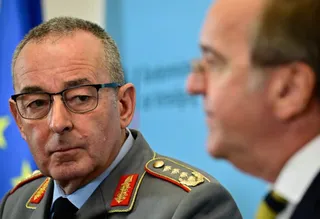 Un bărbat cu ochelari și uniformă militară gri cu epoleți roșii și aurii privește spre un alt bărbat aflat în profil.