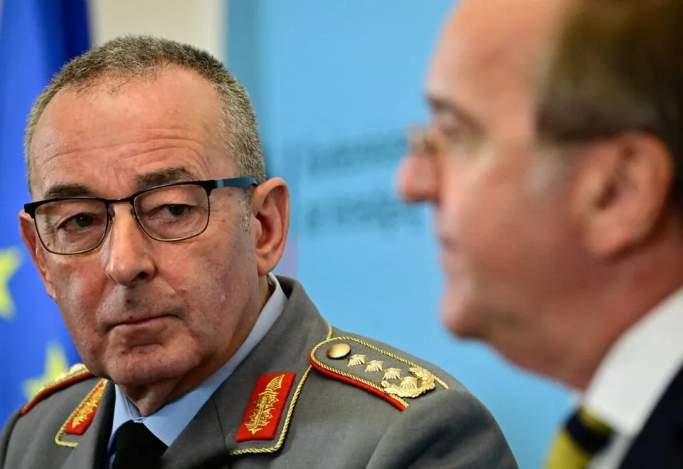 Un bărbat cu ochelari și uniformă militară gri cu epoleți roșii și aurii privește spre un alt bărbat aflat în profil.