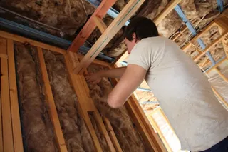 Un bărbat cu tricou alb instalează vată minerală între stâlpii de lemn ai unui perete în interiorul unei construcții.