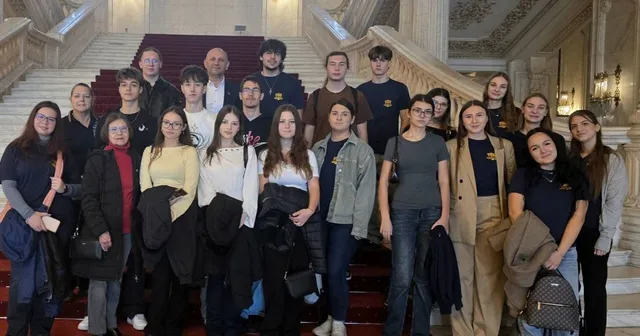 Un grup de tineri stă poziționat pe scările interioare ale unei clădiri mari, purtând haine casual și rucsacuri.