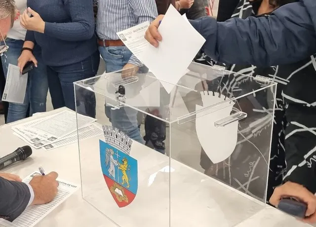 O persoană introduce o foaie de hârtie într-o urnă transparentă de pe o masă albă, lângă documente și un microfon.