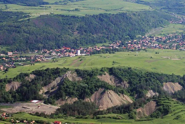 O vedere panoramică asupra unui peisaj format din dealuri verzi, păduri și un sat cu case cu acoperișuri colorate întins pe o vale.