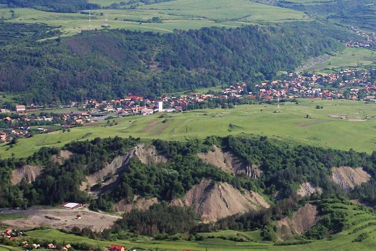 O vedere panoramică asupra unui peisaj format din dealuri verzi, păduri și un sat cu case cu acoperișuri colorate întins pe o vale.