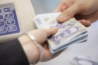 Două mâini țin un teanc de bancnote de o sută de lei lângă un aparat de contabilitate.