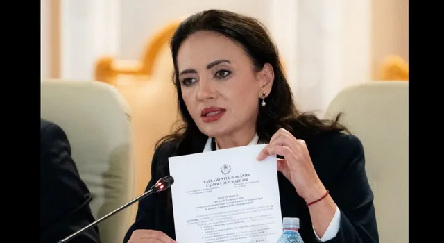 O femeie cu părul închis la culoare, purtând o jachetă neagră și o cămașă albă, ține o foaie de hârtie albă cu text imprimat.