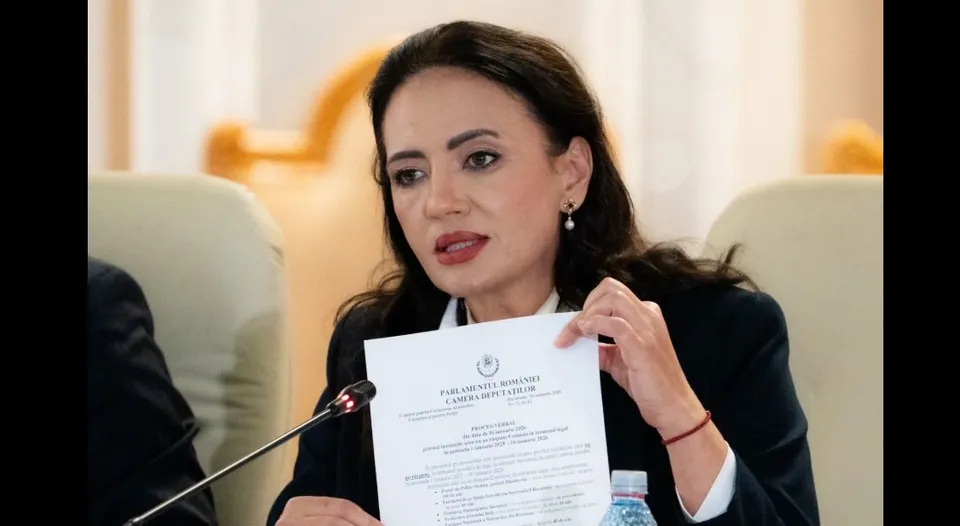 O femeie cu părul închis la culoare, purtând o jachetă neagră și o cămașă albă, ține o foaie de hârtie albă cu text imprimat.