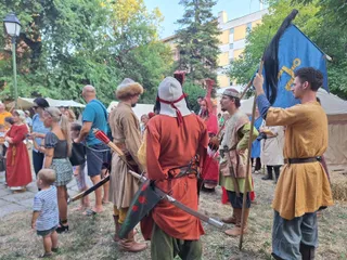 Oamenii poartă haine de epocă și țin steaguri într-un spațiu verde cu copaci și clădiri în fundal.