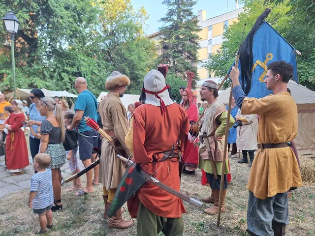 Oamenii poartă haine de epocă și țin steaguri într-un spațiu verde cu copaci și clădiri în fundal.