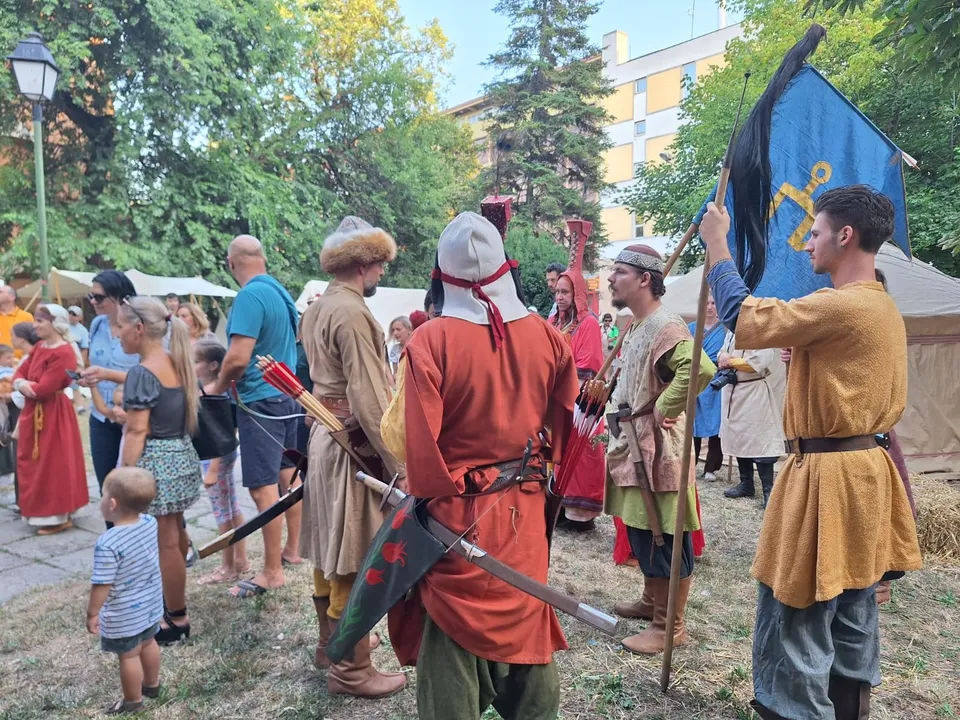 Oamenii poartă haine de epocă și țin steaguri într-un spațiu verde cu copaci și clădiri în fundal.