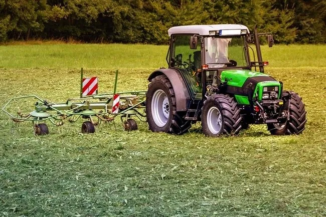 Un tractor verde cu o remorcă agricolă este oprit pe un câmp de iarbă verde sub un fundal de copaci.