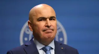 Un bărbat cu capul chel, purtând sacou albastru, cămașă deschisă la culoare și cravată albastră, este surprins într-o prim-plan cu un fundal albastru.