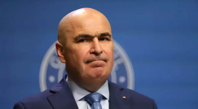 Un bărbat chel, purtând sacou albastru, cămașă albă și cravată albastră, este surprins într-un prim-plan în fața unui fundal albastru cu un logo circular.