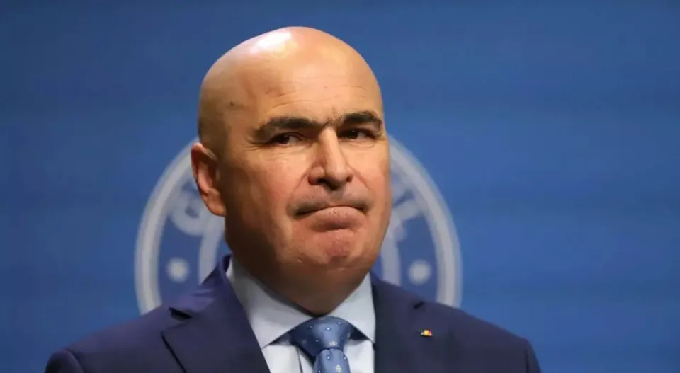 Un bărbat chel, purtând sacou albastru, cămașă albă și cravată albastră, este surprins într-un prim-plan în fața unui fundal albastru cu un logo circular.