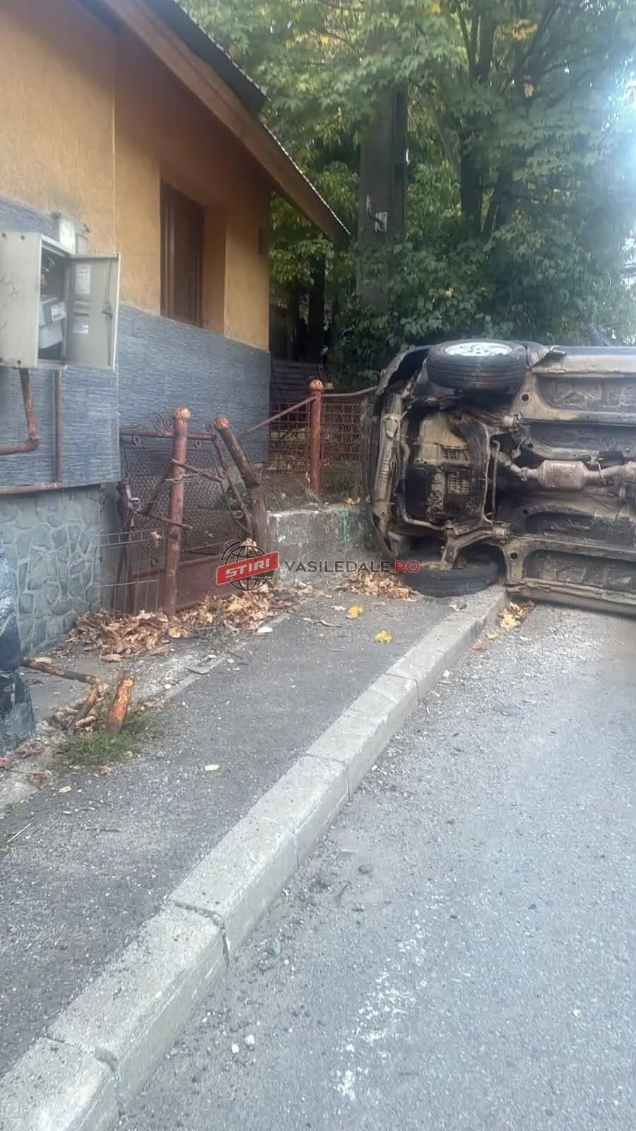 Un autoturism este răsturnat pe o parte lângă un gard metalic și o clădire cu perete galben.