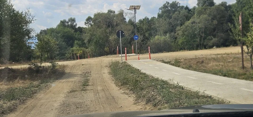 O potecă de pământ și o bandă asfaltată trec prin vegetație sub un cer înnorat, lângă un stâlp cu panou solar.