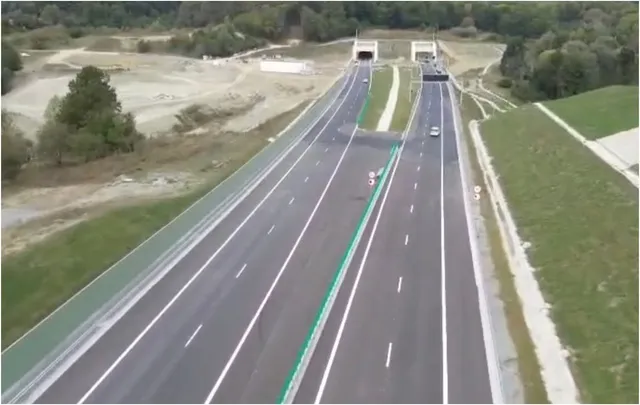 O vedere aeriană prezintă două caraje de autostradă cu marcaje albe și verzi, ducând spre două intrare de tunel în mijlocul unui peisaj de dealuri.