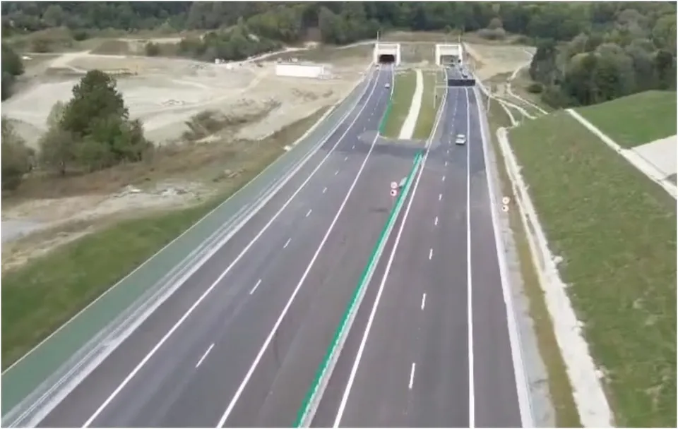 O vedere aeriană prezintă două caraje de autostradă cu marcaje albe și verzi, ducând spre două intrare de tunel în mijlocul unui peisaj de dealuri.