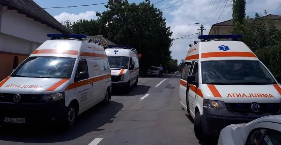 Două ambulanțe albe cu dungi portocalii și un al treilea vehicul similar sunt parcate pe un drum asfaltat lângă clădiri și copaci.