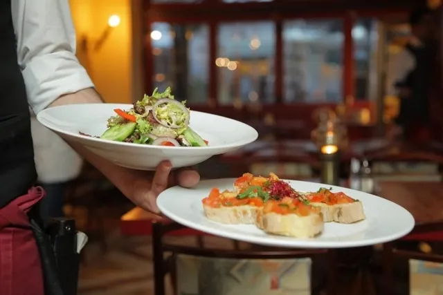 Un chelner poartă două farfurii cu mâncare într-un restaurant, una conținând salată și cealaltă felii de pâine cu topping colorat.