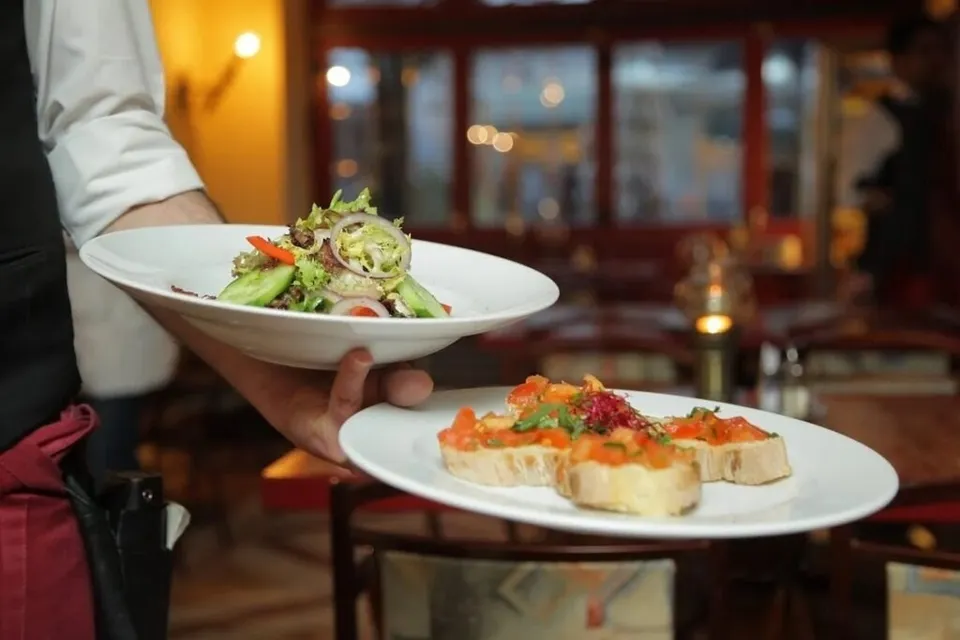 Un chelner poartă două farfurii cu mâncare într-un restaurant, una conținând salată și cealaltă felii de pâine cu topping colorat.