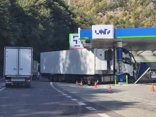 Un autocamion alb cu remorcă frigorifică este poziționat lângă o stație de alimentare, lângă conuri de circulație portocalii pe asfalt.