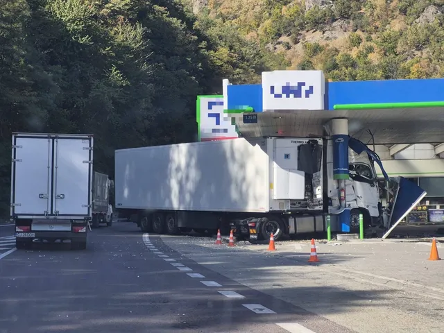 Un autocamion alb cu remorcă frigorifică este poziționat lângă o stație de alimentare, lângă conuri de circulație portocalii pe asfalt.