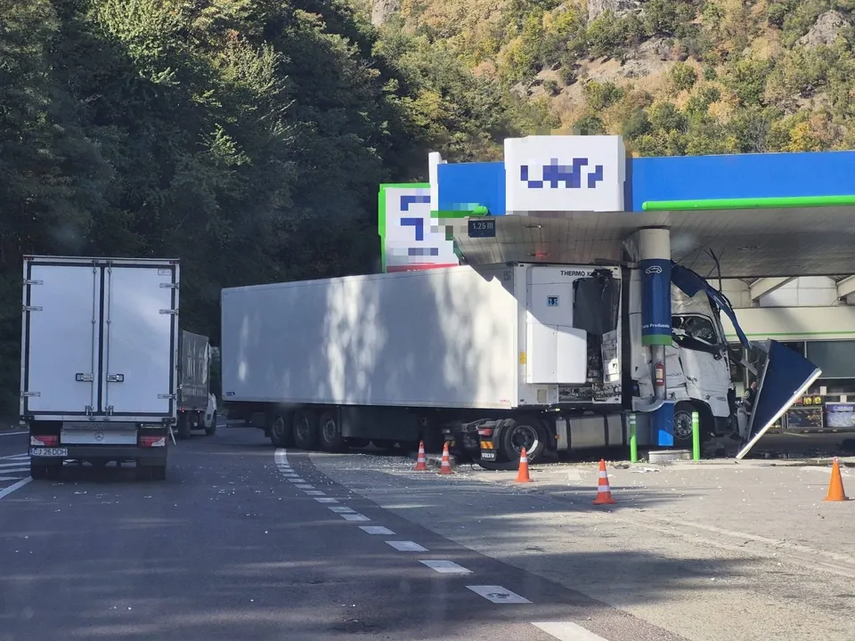Un autocamion alb cu remorcă frigorifică este poziționat lângă o stație de alimentare, lângă conuri de circulație portocalii pe asfalt.