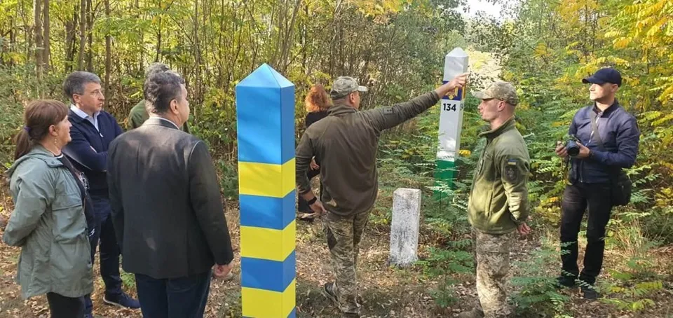 Un grup de persoane stă într-o pădure lângă un stâlp alb cu marcaje verzi și un stâlp albastru și galben.