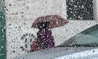 O persoană cu o umbrelă purtată sub ploaie este vizibilă prin picăturile de apă de pe o suprafață transparentă.