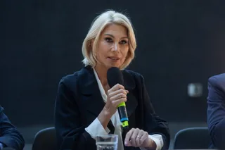 O femeie cu părul blond, purtând o jachetă neagră peste o cămașă albă, ține un microfon negru cu detalii verzi în mână.