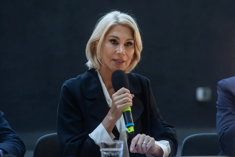 O femeie cu părul blond, purtând o jachetă neagră peste o cămașă albă, ține un microfon negru cu detalii verzi în mână.
