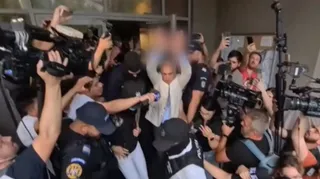 Un bărbat cu o vestă albă este înconjurat de persoane cu camere video, microfoane și polițiști în uniformă întunecată.