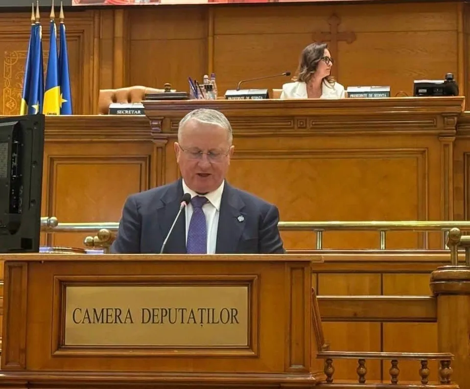 Un bărbat cu ochelari și cravată purpuresie stă în spatele unui pupitru de lemn pe care scrie Camera Deputaților.