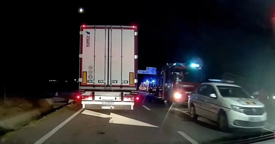 Un camion alb și o mașină de poliție cu luminile aprinse sunt vizibile pe o stradă asfaltată pe timp de noapte.