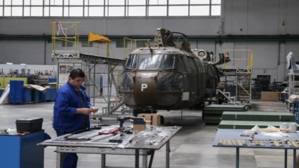 Un bărbat într-un costum de lucru albastru stă lângă o masă de lucru într-un hangar lângă un elicopter mare de culoare închisă.