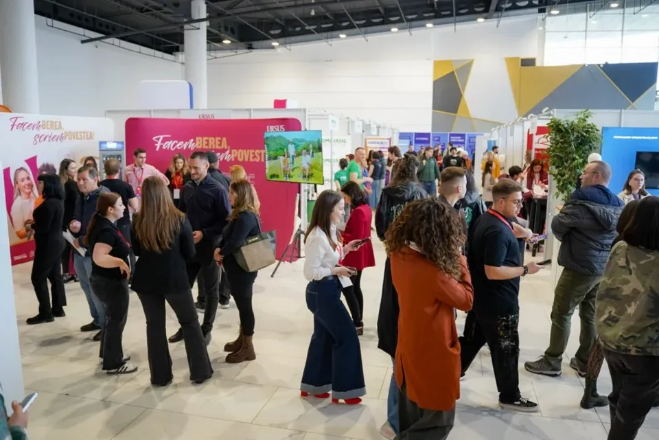 Oamenii stau în picioare într-o sală de evenimente cu standuri de prezentare și panouri colorate în fundal.