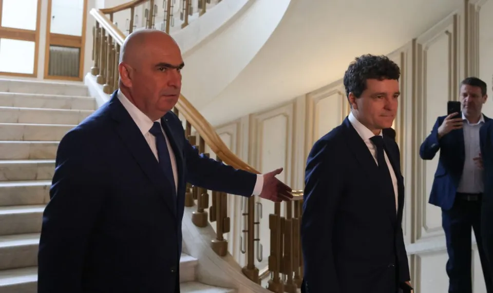 Trei bărbați în costume închise descendo o scară interioară cu balustradă metalică și mâner de lemn.