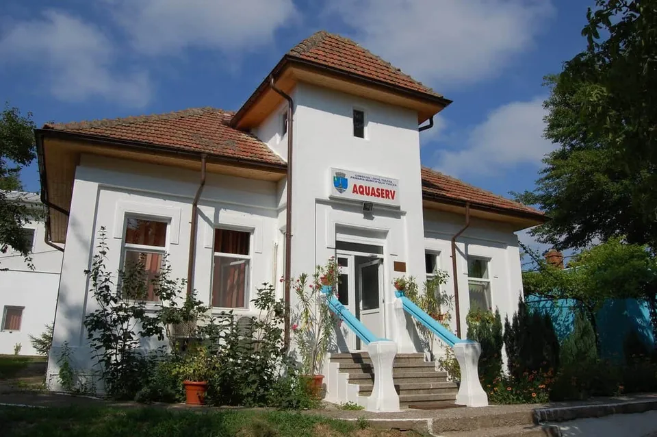 O clădire albă cu acoperiș de țiglă roșie, un logo albastru și roșu pe față, scări exterioare cu balustrade albastre, și vegetație verde în jur.