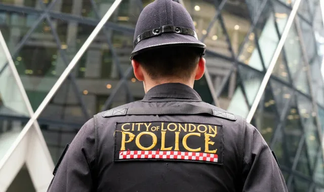 Un polițist cu spatele la cameră poartă o uniformă neagră pe care este aplicat un patch cu inscripția CITY OF LONDON POLICE.