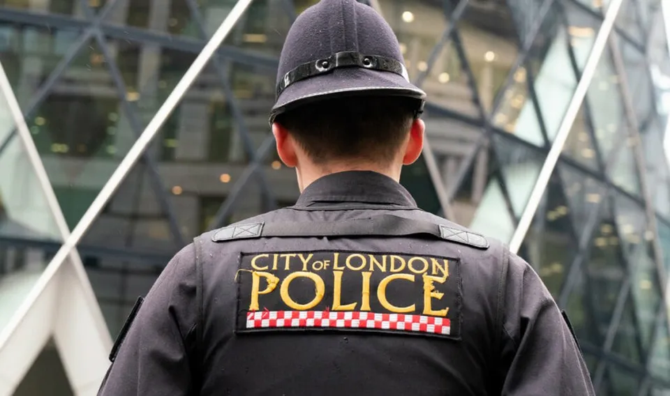 Un polițist cu spatele la cameră poartă o uniformă neagră pe care este aplicat un patch cu inscripția CITY OF LONDON POLICE.