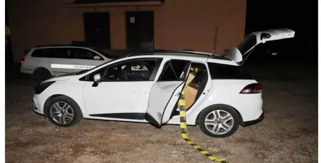 Un autoturism alb cu portbagajul și portierea din spate deschise este parcat pe un teren pietruios lângă o clădire în timpul nopții.