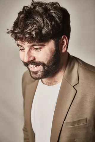 Un bărbat cu părul închis la culoare și barbă poartă un sacou maro peste un tricou alb, privind în jos cu un zâmbet ușor.