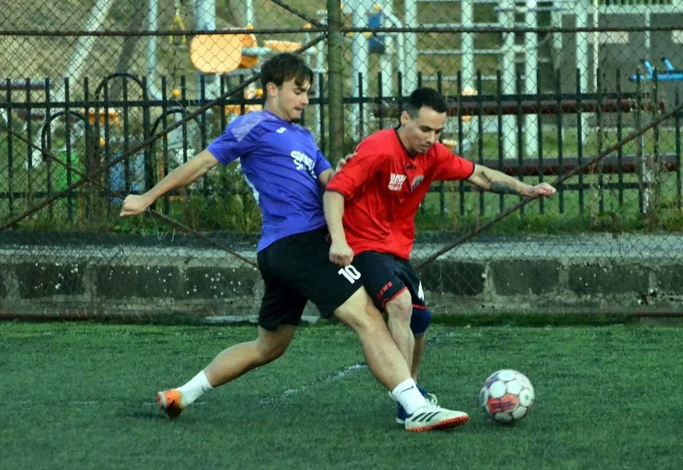 Un jucător în echipament albastru și unul în echipament roșu dispută un balon pe un teren de fotbal cu iarbă sintetică.