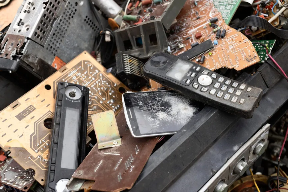 Un grup de componente electronice, plăci de circuit imprimat și un telefon mobil cu ecranul spart sunt depozitate într-un amestec de deșeuri.