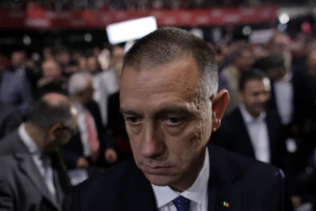 Un bărbat cu părul scurt și costum în prim-plan privește în jos, în timp ce o mulțime de oameni stă în fundalul neclar.