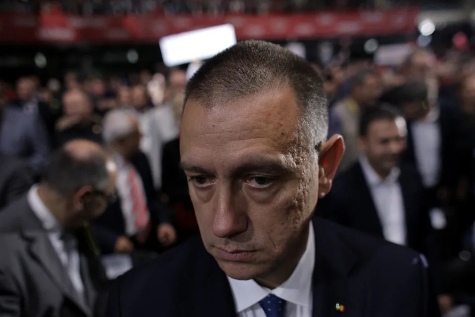 Un bărbat cu părul scurt și costum în prim-plan privește în jos, în timp ce o mulțime de oameni stă în fundalul neclar.