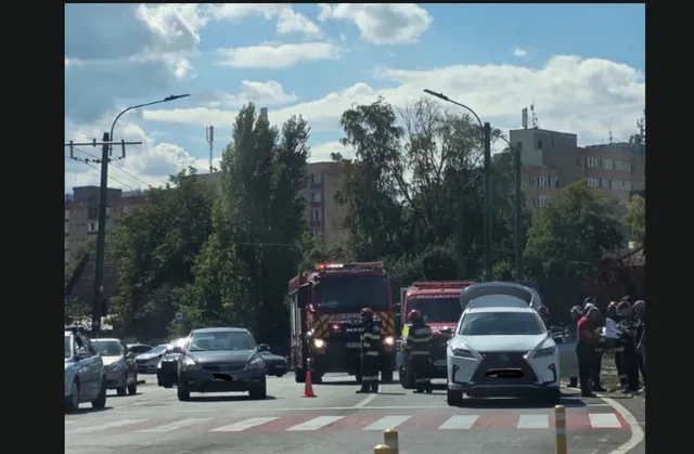 O intersecție aglomerată cu mașini, un camion de pompieri, și mai mulți oameni, sugerând un accident rutier. Cerul este parțial înnorat.